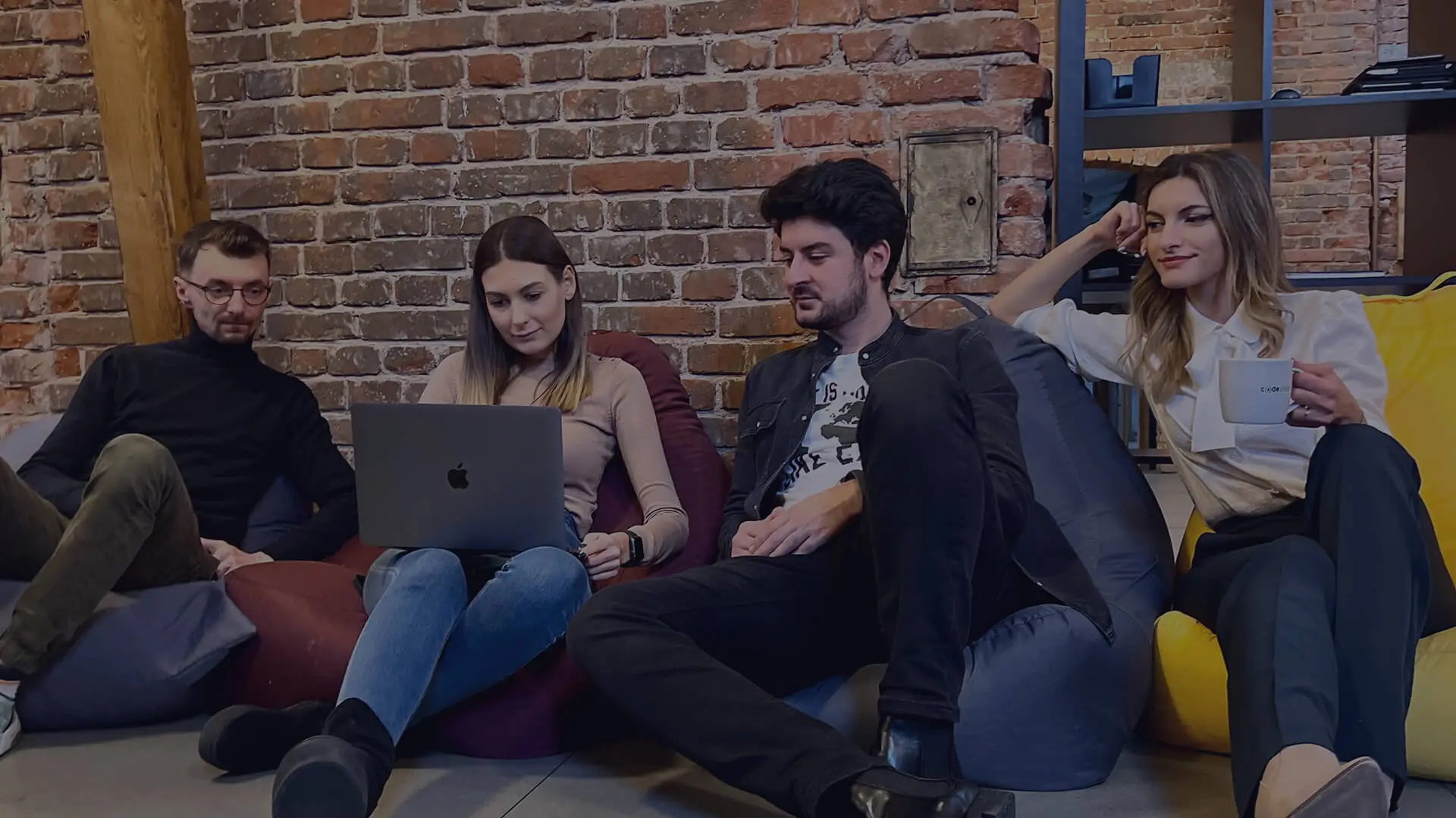Un grup de programatori și designeri care lucrează împreună la un laptop, așezați pe beanbaguri.