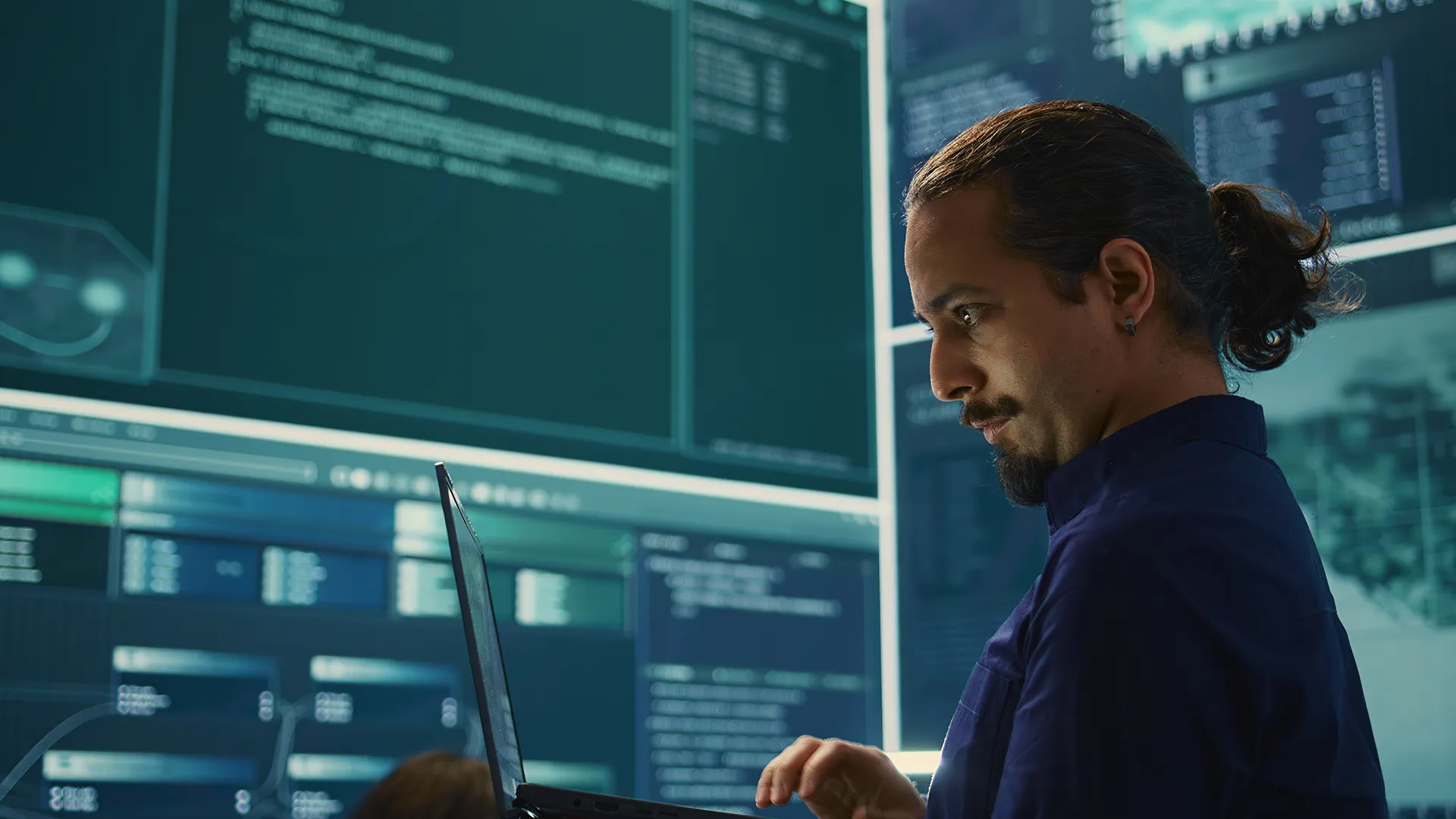 Software engineer working on a laptop in front of multiple screens displaying code and data